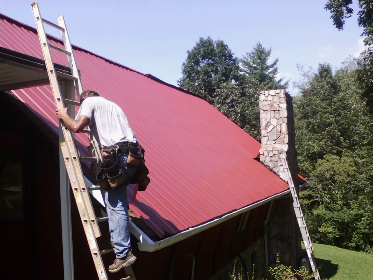 Roofer on a ladder beside a red metal roof and stone chimney — Soffit & Fascia Repair by Roofing Brothers in Peaks Island